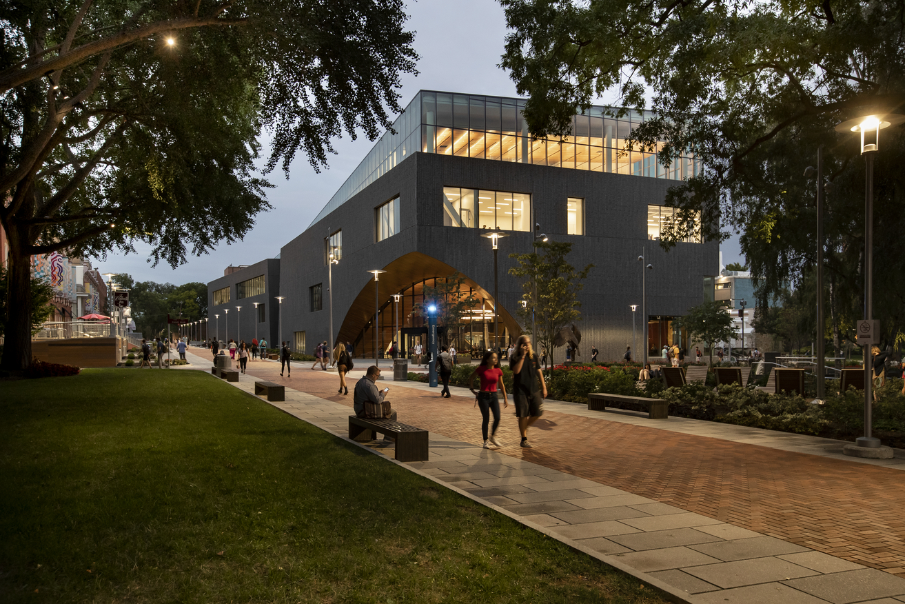 Charles Library at night. People walk on a brick sidewalk among trees and other greenery.