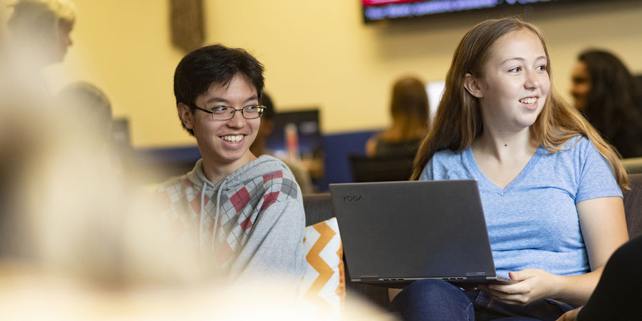 Two students sit in a lounge with their laptops, looking to the right and smiling.
