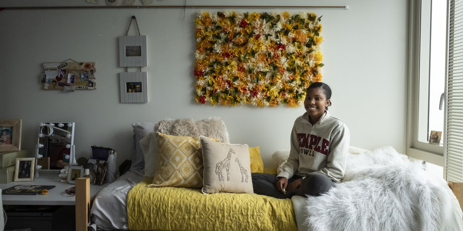 A smiling Temple student sits on their bed in their dorm room.