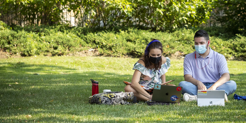 two students wearing face coverings studying in the grass.