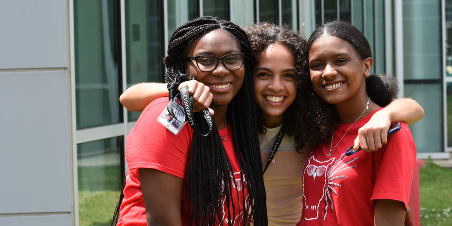 Three female pre-college students stand with their arms around each other, posing for a photo.