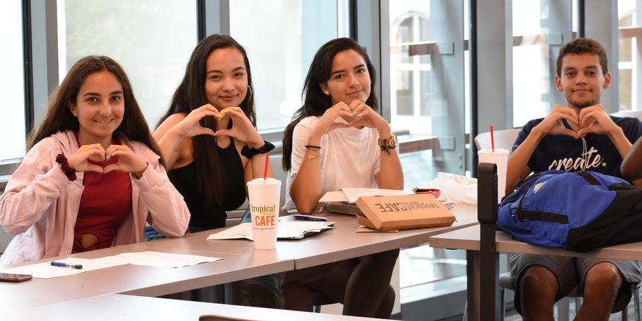 Group of male and female pre-college students sit together in a classroom and hold up their hands in the shape of hearts.
