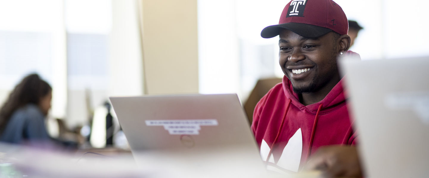 student wearing a baseball cap and smiling while working on a laptop