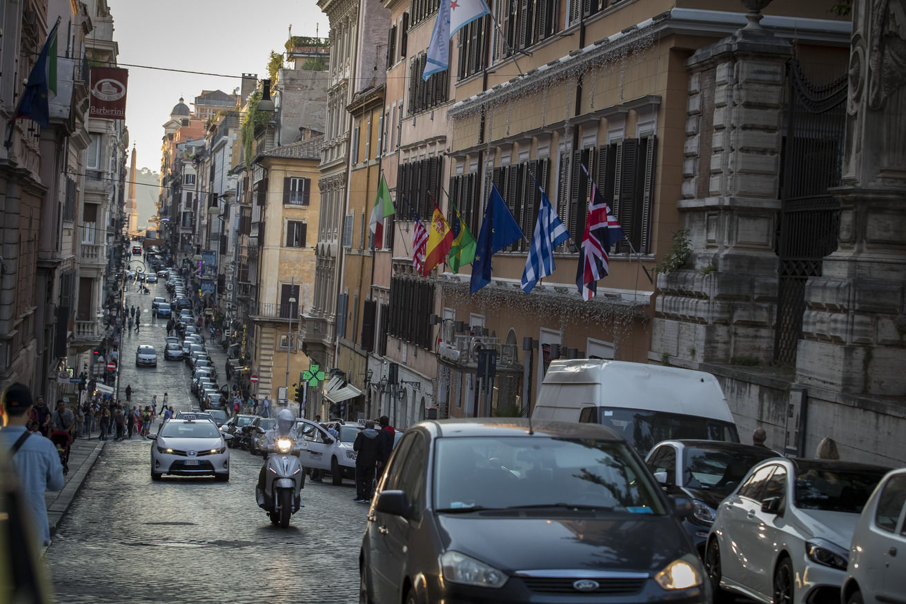 A busy cobblestone road in Rome filled with cars, motorbikes and pedestrians. 