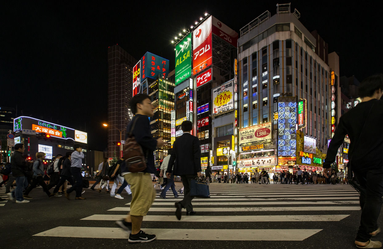 People crossing a busy intersection in a trendy neighborhood in central Tokyo with many brightly lit signs on the facades of the buildings. 