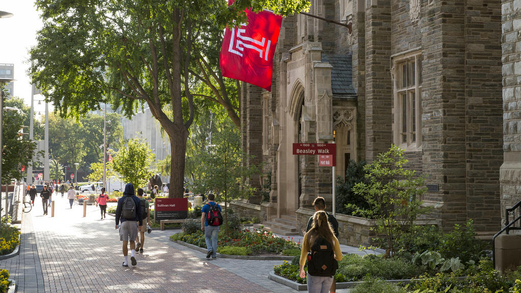College students walk across campus on a sunny day with Temple University flag