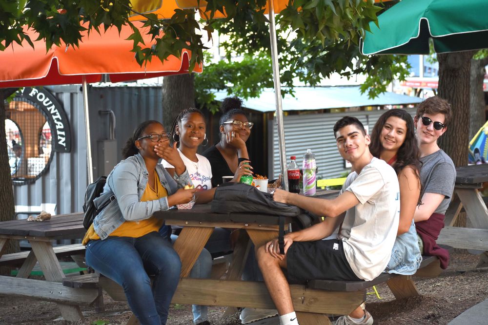 A group of pre-college students and mentors sit at a wooden picnic table at Spruce Street Harbor Park.